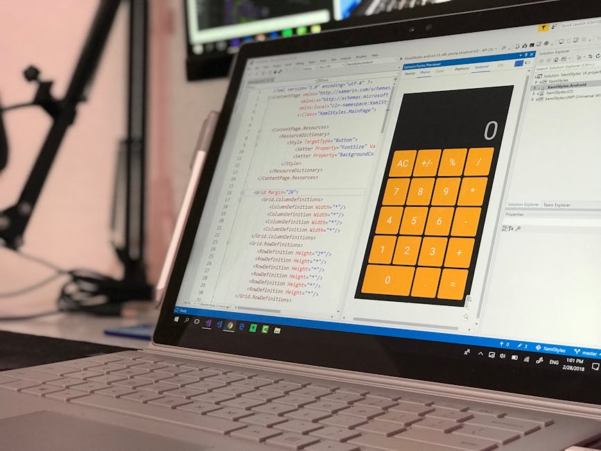 Close-up of a laptop computer on a desk displaying a web development code editor alongside a large calculator application with orange buttons and a zero key, set in an indoor workspace with a blurred