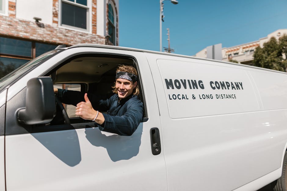 A white moving van parked in front of a two-Storey residential building with a brick and white exterior, during daylight hours under a clear blue sky. The van has the words 'MOVING COMPANY' and 'LOCAL